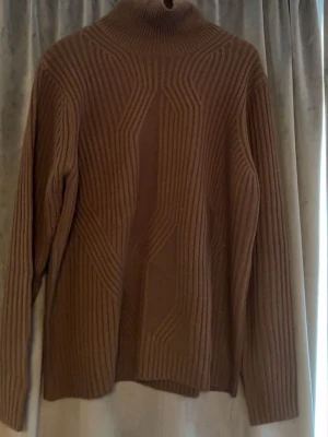 Brun ribbstickad polotröja - Säljer en brun ribbstickad polotröja med lång ärm och hög krage. Tröjan har ett coolt geometriskt mönster framtill och är gjord i ett mjukt, tjockt material som håller dig varm. Perfekt för dig som gillar stilrena och trendiga plagg.