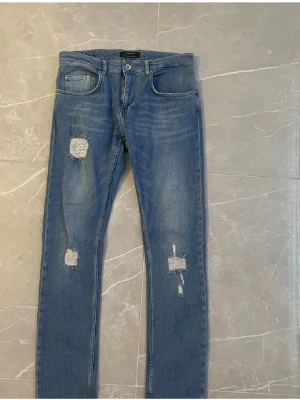 Blå slitna skinny jeans - Säljer ett par blå skinny jeans med slitningar och hål på båda benen. Jeansen har klassisk femficksdesign, normalhög midja och dragkedja med knapp. Perfekta för en avslappnad och trendig look.
