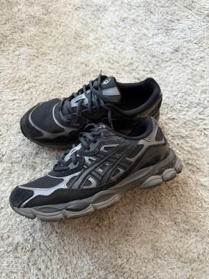 Svarta och grå Asics sneakers med meshdetaljer - Snygga sneakers i svart och grått med meshpaneler och detaljer i mockaimitation. Skorna har en chunky sula och klassisk snörning. Perfekta för dig som gillar sportig stil och vill ha bekväma skor till vardags.