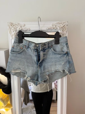 Ljusa jeansshorts med råa kanter - Säljer ett par ljusblå jeansshorts med hög midja och slitna, fransiga kanter. Klassisk femficksmodell med dragkedja och knapp framtill. Perfekta för sommaren och ger en avslappnad streetstyle-look.