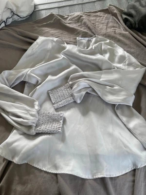 White Satin Blouse with Smocked Cuffs - A long-sleeved, white satin blouse featuring wide, smocked cuffs. The blouse has a soft, shiny texture.