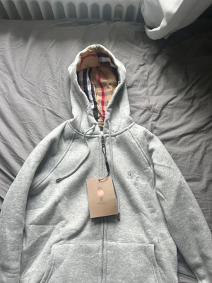 Grå hoodie från Burberry med huva - Snygg grå zip-hoodie från Burberry med klassiskt rutigt foder i huvan och broderad logga på bröstet. Hoodien har dragsko i huvan och stora fickor framtill. Tröjan är liten i storlek