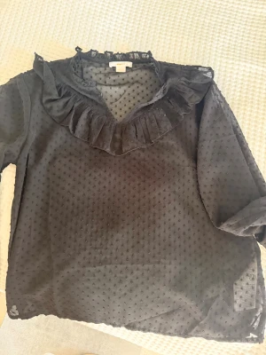 Svart blus med volang från Gina Tricot - Snygg svart blus från Gina Tricot med volangdetalj runt halsen och diskret prickigt mönster i tyget. Blusen är gjord i ett tunt, transparent material och har långa ärmar. Perfekt för dig som gillar trendiga detaljer och en luftig känsla.