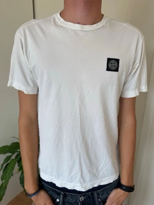 Vit Stone Island t-shirt - Vit t-shirt från Stone Island med klassisk svart patch på bröstet. T-shirten har rund hals och korta ärmar, tillverkad i mjuk bomull för en skön och avslappnad look. Perfekt till jeans eller shorts för en clean stil.