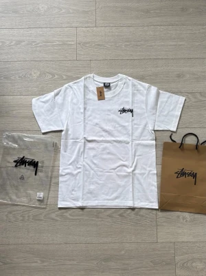 Vit Stüssy t-shirt med tärningsmotiv - Vit t-shirt från Stüssy med svart logga på bröstet och stort tryck av två rosa tärningar samt logga på ryggen. 
