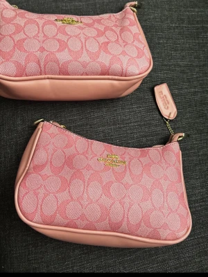 Rosa handväska från Coach - Säljer en snygg rosa handväska från Coach med det klassiska C-mönstret i tyg och detaljer i ljusrosa läder. Väskan har guldfärgade metalldetaljer och en kedjerem som ger en trendig look. Perfekt för dig som vill ha något unikt och stilrent.