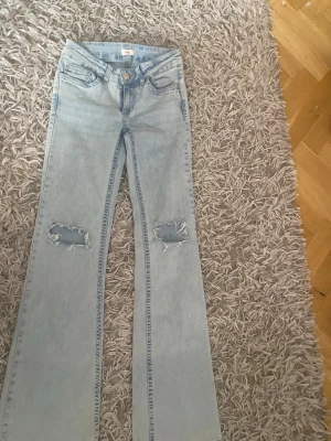 nelly jeans - Säljer ett par ljusblå bootcut jeans med slitna hål på knäna och broderade detaljer på bakfickorna. Jeansen har klassisk femficksmodell och är i ett mjukt bomullsmaterial. Perfekta för en avslappnad och trendig look.