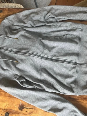 Grå zip hoodie från essentials - Snygg grå hoodie från ESSENTIALS med dragkedja. Klassisk modell med huva och justerbara snören. Tillverkad i mjukt sweatshirtmaterial som är skönt mot huden. Perfekt för en avslappnad stil.