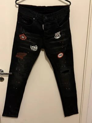 Dsquared2 Slim Jean – Svarta jeans med patchar (Strl 46) - Säljer ett par äkta Dsquared2 Slim Jeans i svart (Modell: S71LB0589) från 2022 års kollektion. Jeansen har snygga detaljer som "Route 64" och "DSQ2" patchar, färgstänk (paint splatter) samt slitningar. Tillverkade i Italien. 