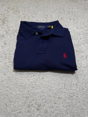 Marinblå pikétröja från Polo Ralph Lauren - Klassisk marinblå pikétröja från Polo Ralph Lauren med röd broderad logga på bröstet. Tröjan har krage, knappar framtill och korta ärmar. Perfekt till en avslappnad men ändå stilren look.