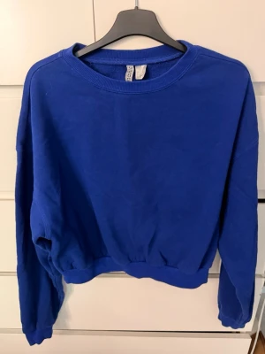 sweatshirt - En enkel och stilren blå sweatshirt från Divided. Tröjan har rund halsringning, ribbade muddar vid ärmslut och midja samt en avslappnad passform. Tillverkad i mjukt bomullsmaterial som är skönt mot huden. Perfekt för en chill och casual look.