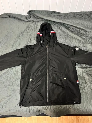 Moncler vindjacka - Snygg svart vindjacka med huva och dragkedja framtill. Jackan har två fickor med tryckknappar och detaljer i rött, vitt och blått på huvan och sidan. Liten rund patch på ena ärmen. Perfekt för blåsiga dagar och enkel att matcha.