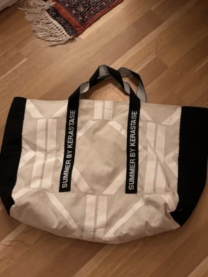 Stor shoppingväska i tyg - Säljer en stor och rymlig shoppingväska i beige och svart tyg med grafiskt mönster. Väskan har svarta handtag med texten 'SUMMER BY KERASTASE' i vitt. Perfekt för att bära med sig allt man behöver till stranden eller stan.