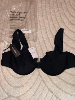 Svart bikiniöverdel PrettyLittleThing - Stilren svart bikiniöverdel från PrettyLittleThing med breda axelband och snygg metallspänne framtill. Bikinin har en enkel och klassisk design som passar perfekt till sommaren. Materialet känns mjukt och följsamt. Helt oanvänd 