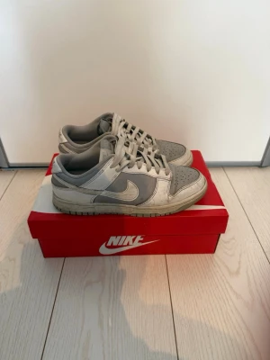 Nike Dunk Low grå/vita sneakers - Nike Dunk Low sneakers i grått och vitt, storlek 40! Bra skick, inga större skavanker/ slitningar i alla fall inget som stör vid användning! 