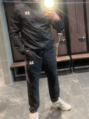 Svart träningsset från Under Armour - Säljer ett svart träningsset från Under Armour med matchande jacka och byxor. Setet har vita loggor på både jacka och byxa, dragkedja på jackan och huva. Perfekt för gymmet eller löprundan. 