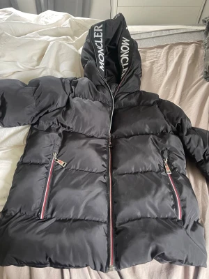 Svart dunjacka ( inte äkta moncler)  - Snygg svart dunjacka med stor huva och tydlig logga på huvan. Jackan har två framfickor med dragkedjor och röda/vita detaljer längs dragkedjorna. Puffig modell som håller dig varm under kalla dagar. Perfekt för vintern och riktigt stilren. Den är i bra skick, dock kan man se att den är använd vid tillexempel dragkedjan ( se bild 3). 