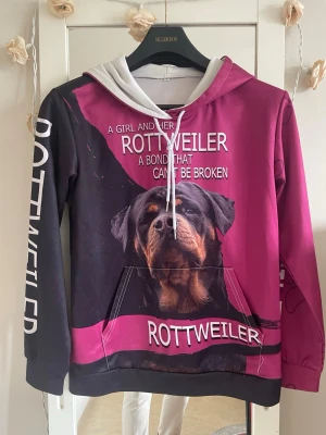 Rosa och svart hoodie med Rottweiler - Unik hoodie i rosa och svart med stor Rottweiler-tryck både fram och bak. Texten 'A girl and her Rottweiler, a bond that can't be broken' på framsidan och 'ROTTWEILER' på ärmen. Har huva med vita snören och magficka. Perfekt för hundälskare!storleken är S men som medium går också 