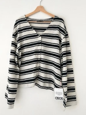 Randig stickad kofta från ASOS DESIGN - Snygg kofta från ASOS DESIGN i vitt och svart med randigt mönster. Koftan har v-ringning, knappar framtill och är stickad i mjukt material. Perfekt för lager-på-lager och chill vardagsstil.