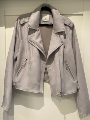 Bikerjacka i mockaimitation - Ljusgrå kort jacka i bikermodell, mockaimitation. Är ganska stretchig i modellen. 