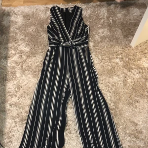 H&M black jumpsuit with white stripes  - Svart jumpsuit från H&M i storlek 38. Plagget har endast använts 1 gång och är i nyskick. Den har en cut-out front med ett insytt bälte som man kan knyta runt midjan med en drag kedja i bak. 