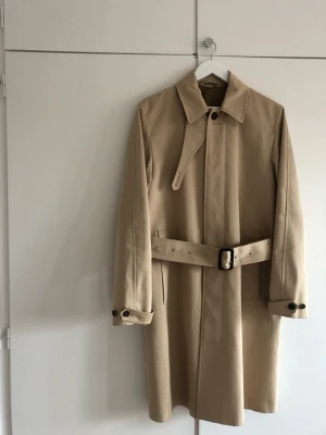 Tiger of Sweden trenchcoat strl 48 - Trenchcoat i klassisk rak passform med bälte i midjan. Rocken ner till knäna om man är runt 175 cm lång. Rocken är även vattenavvisande 