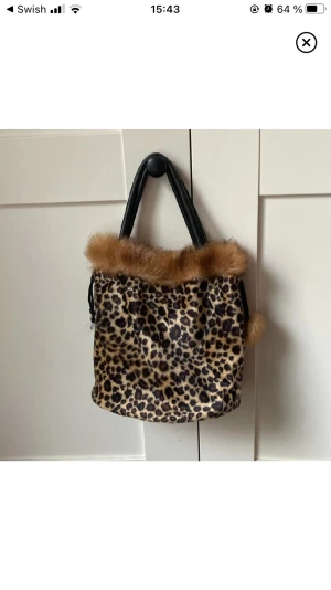 Leopard väska - Väska med leopard/cheetah mönster med faux furr. Den går att stänga med tråden på vardera sida. Har några defekter på undersidan och tråden. Mått: höjd: 20cm längd 19cm, bredd/djup: 12cm.