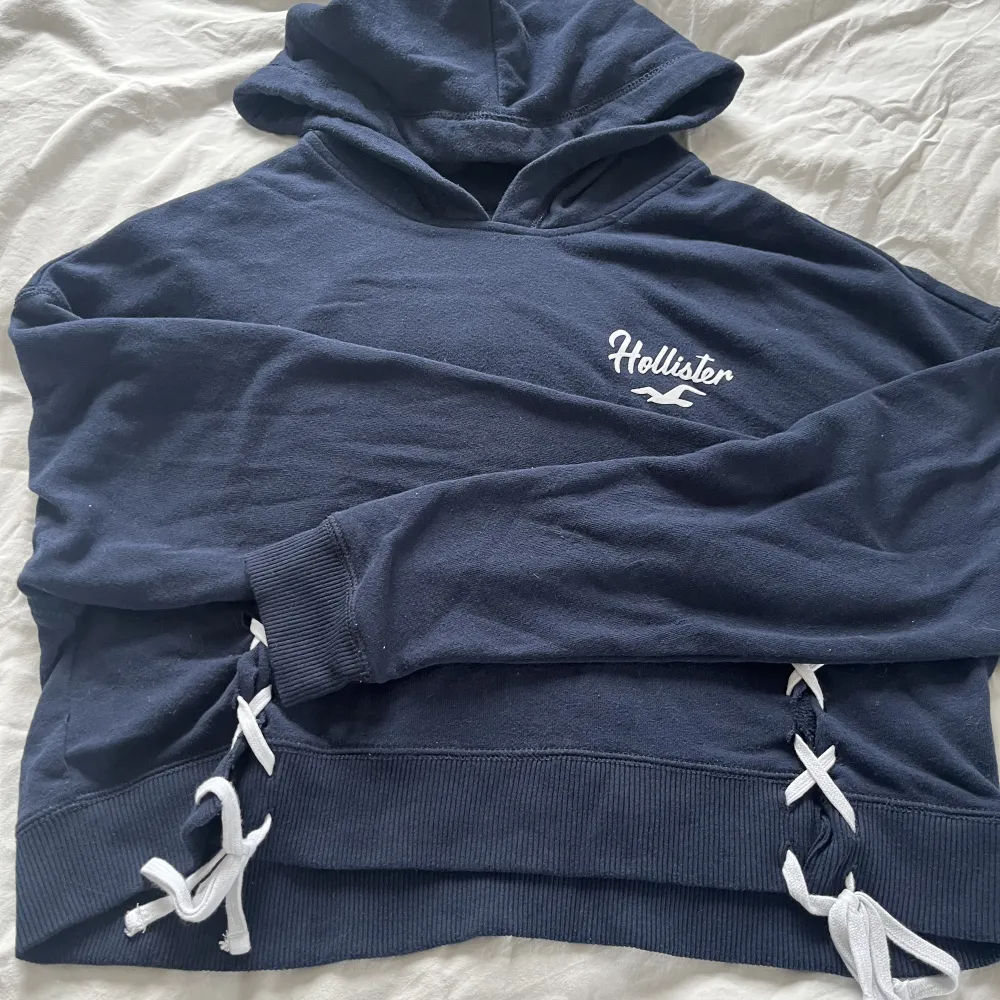 En marinblå hoodie från hollister med coola detaljer. Fint tryck på ryggen. Den är xs, men passar fint på mig som brukar ha m med bara att den är lite mer croppad då.🥰. Hoodies.