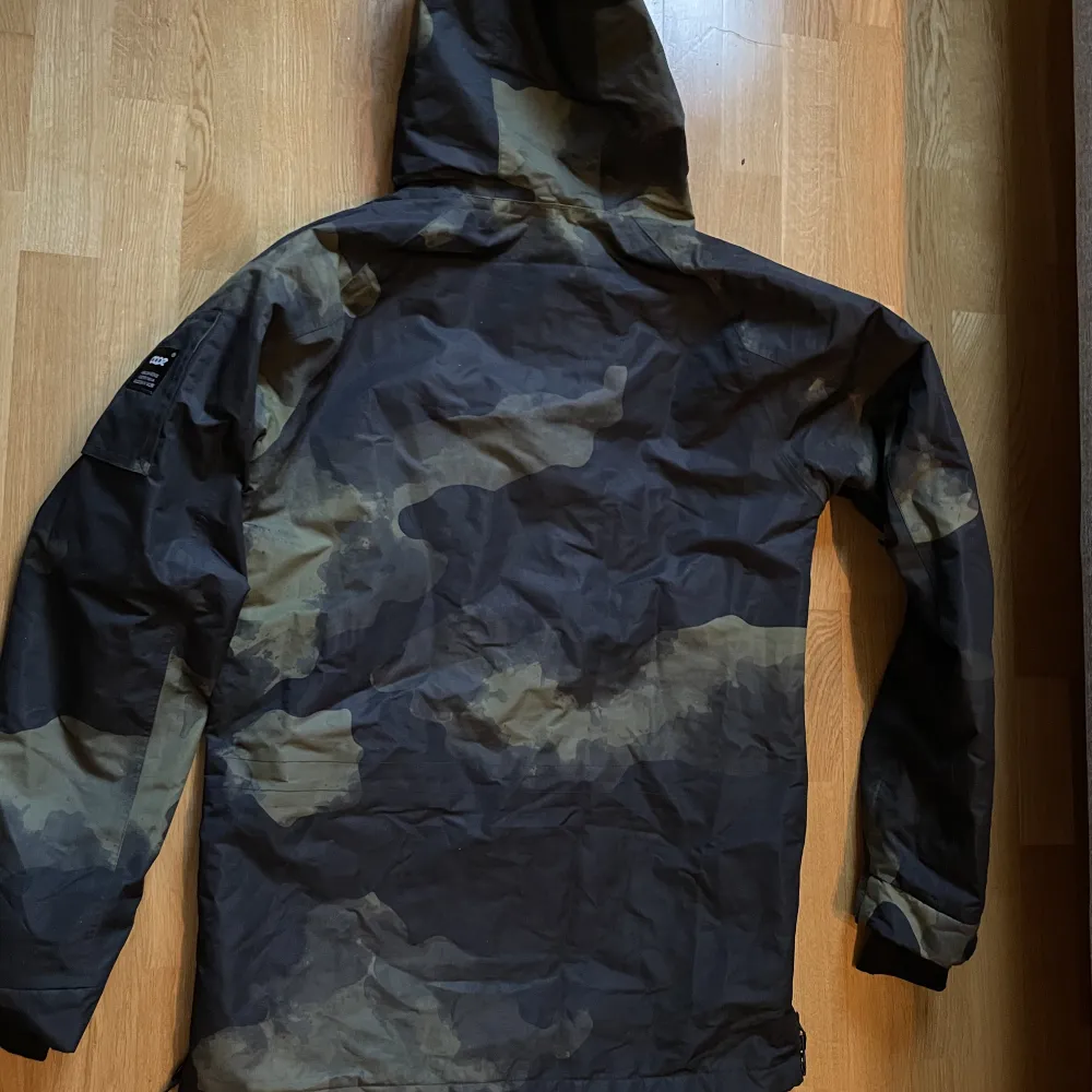Anorak skidjacka från märke Dope. Militärmönster. Två st fickor på magen. En ficka för liftkort på ena ärmen. Dragkedjor på sidorna för ventilation. Luva som går att få över skidhjälm. Använd 1 vecka i fjällen. Inget tecken på slitage. . Takit.