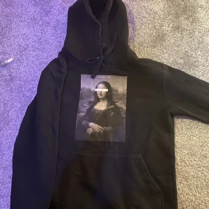 Mona Lisa hoodie  - En hoodie med ett tryck med Mona Lisa köpt från The cool elephant. Har växt ur den så vill bara bli av med den! Den är inte använd så mycket, om man vill ha mer detaljer kan man kontakta mig! Trycket är ytterst lite påverkat som knappt syns. 