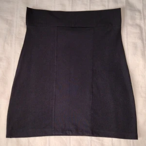 Svart kort kjol med knytband, storlek XS/S - Svart kort kjol med knytning i ryggen. Har tidigare varit en klänning och överdelen har klippts bort, vilket bara syns spår av på insidan. Väldigt mjuk och elastisk. Passar storlek XS/S skulle jag säga. Sparsamt använd.