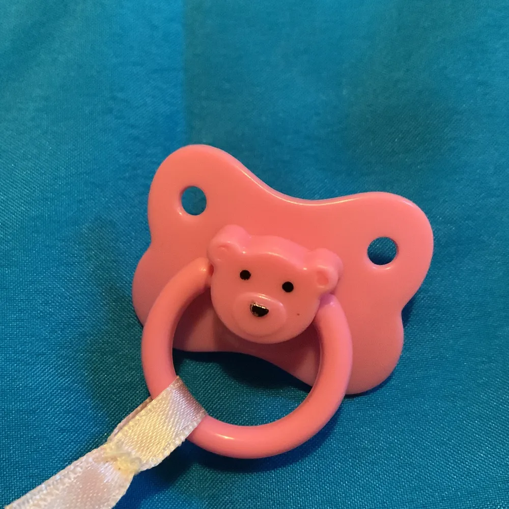 Det är en bebis med en rosa overall. Den har också en rosa napp med björnform. . Övrigt.