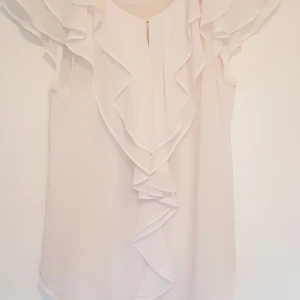 Vit blus - Söt vit blus i skirt tyg. Litet knäppe på framsidan som går att ha öppet eller stängt. Väl använd, men fortfarande i gott skick. Pris exkl frakt. Hämtas på Lilla essingen alt mötas upp på Fridhemsplan.