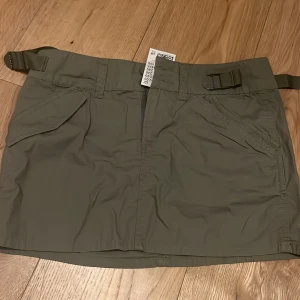 Cargo skirt - En supercool cargokjol i färgen militärgrön. Jättefina detaljer, med justerbara band runt midjan. Aldrig använd, och lappen sitter fortfarande på.  