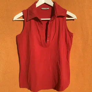 Rött linne med krage och v-urringning med knappar. I bomullsmaterial med lite stretch. Tajt passform. Om du har frågor eller vill se fler bilder är det bara att skicka iväg ett meddelande:)
