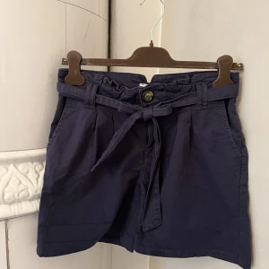 Mörkblå highwaste shorts - Mörkblå/marinblå högmidjade shorts i tyg, med knytskärp och knapp. Fickor fram och bak. Strl Xs/S. Går att vika upp om man vill ha kortare ben. Använt skick.