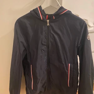 Mörkblå vindjacka från Moncler - Snygg svart vindjacka från Moncler. Självklart är den äkta, köpt på Moncler hemsidan i storlek 14y. Nypris på den ligger på 6050kr men jag säljer den för 1900kr då jag inte längre har något som medföljer. Pris kan diskuteras vid snabb affär!!