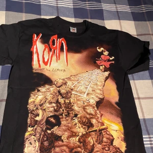 Korn t-shirt Fruit of the Loom - Cool svart t-shirt med Korn-tryck fram och bak. Framsidan har ett färgglatt motiv med texten 'Follow the Leader'. Perfekt för fans av bandet!