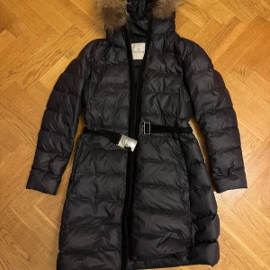 Svart dunjacka från Moncler - Snygg svart dunjacka från Moncler med pälskantad huva och bälte i midjan. Jackan har en lång design med dragkedja framtill och är perfekt för kalla dagar.