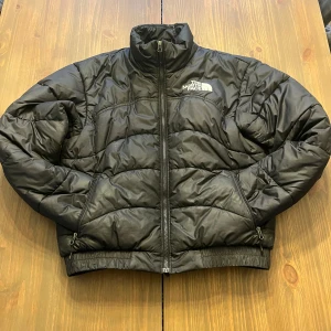 Svart pufferjacka från The North Face - Säljer en svart pufferjacka från The North Face. Jackan har en dragkedja framtill och två sidofickor med dragkedjor. Den är quiltad och har en hög krage för extra värme. Perfekt för kyliga dagar.