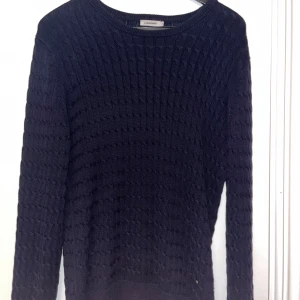 Mörkblå stickad tröja från J.Lindeberg  - Snygg mörkblå stickad tröja från J.Lindeberg med ett klassiskt kabelstickat mönster. Tröjan har rund hals och långa ärmar, perfekt för kyligare dagar. Den är både stilren och bekväm.