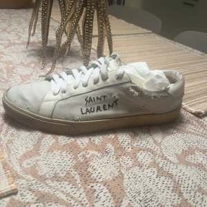 Vita sneakers från Saint Laurent - Säljer ett par vita sneakers från Saint Laurent klassisk design med snörning och en något sliten look som ger dem karaktär. Skorna är helt okej skick❤️pris går att diskutera! Tar Gjärna byten😍ALLT og ingår! Självfallet äkta! 