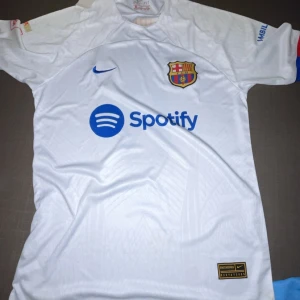 Vit FC Barcelona fotbollströja - Snygg vit FC Barcelona tröja med korta ärmar och klubbens emblem på bröstet. Har sponsorloggor från Spotify och UNHCR. Perfekt för fans som vill visa sitt stöd.