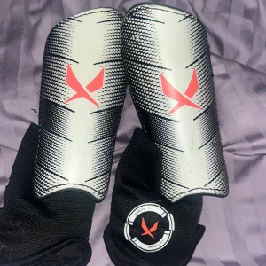 Svarta fotbollsskydd med röd logotyp - Säljer ett par svarta fotbollsskydd med en röd logotyp på framsidan. Skydden har en vit och svart design med elastiska band för att hålla dem på plats. Perfekta för fotbollsträning eller match.