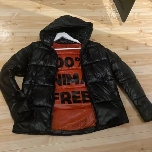 Svart pufferjacka från Save the Duck - Säljer en svart pufferjacka från Save the Duck med huva och dragkedja. Insidan är orange med texten '100% Animal Free'. Perfekt för kyliga dagar. Skicket är 9/10✅ Inga tecken på hål eller liknande