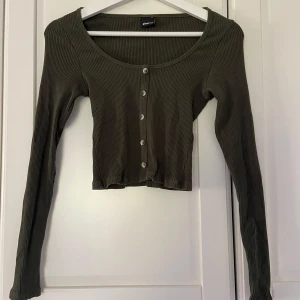 Olivgrön ribbad topp från Gina Tricot - Snygg olivgrön ribbad topp från Gina Tricot med knappar framtill. Den är croppad och har långa ärmar, perfekt för en trendig look. Passar bra till både jeans och kjol.