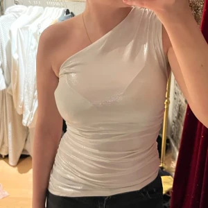 Vit oneshoulder topp - Snygg vit oneshoulder topp med glittrig yta. Perfekt för en kväll ute eller en speciell tillställning. Toppen har en tight passform som framhäver figuren.