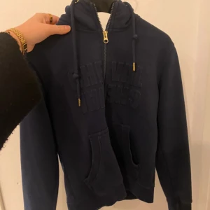 Mörkblå hoodie med dragkedja - Säljer en mörkblå hoodie med dragkedja och justerbar huva. Tröjan har en broderad text på framsidan och praktiska fickor. Perfekt för en avslappnad stil. Köpte på hööks så kan användas som en rid tröja 