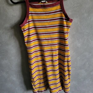 Randig klänning från Monki - Säljer en färgglad randig klänning från Monki i storlek XXS. Klänningen har ett mönster i gult, vinrött och vitt med en ribbad struktur. Den är ärmlös och perfekt för varma dagar.