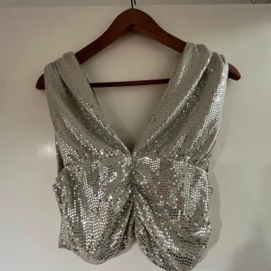 Silvrig glittrig topp - Snygg silvrig topp med glittriga paljetter och djup v-ringning. Toppen är croppad och har en åtsittande passform med rynkade detaljer framtill. Perfekt för nyår